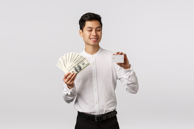 Finance, economy and business concept. Handsome asian businessman in formal outfit, holding cash and credit card, looking at banking payment method with pleased smile, spend money