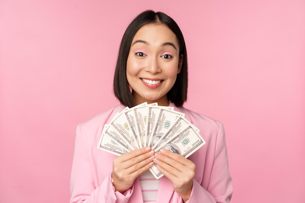 Finance microcredit and people concept Happy smiling asian businesswoman showing dollars money standing in suit against pink background