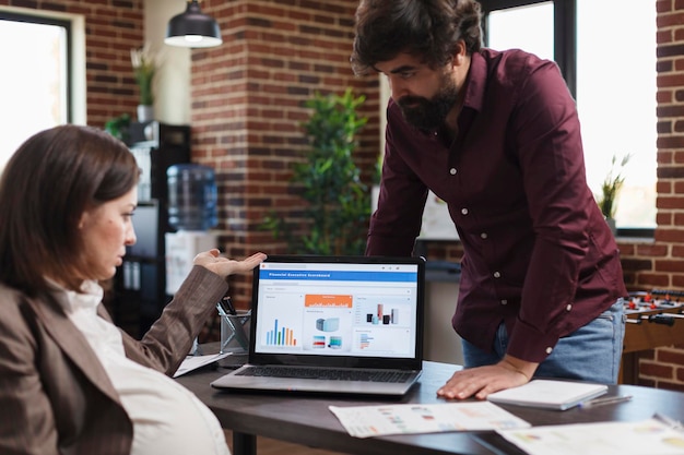 Financial agency office worker informing finance department manager about poor project strategy. Pregnant woman questioning data charts and business analytics results while sitting at office desk.