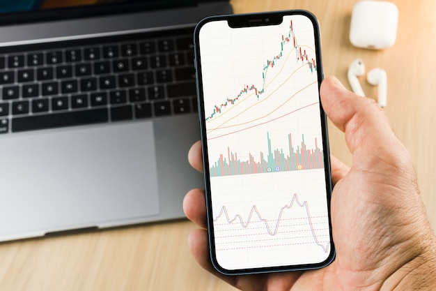 その横にコンピューターと木製の背景のスマートフォン画面上の金融株式市場のグラフ。証券取引所。
