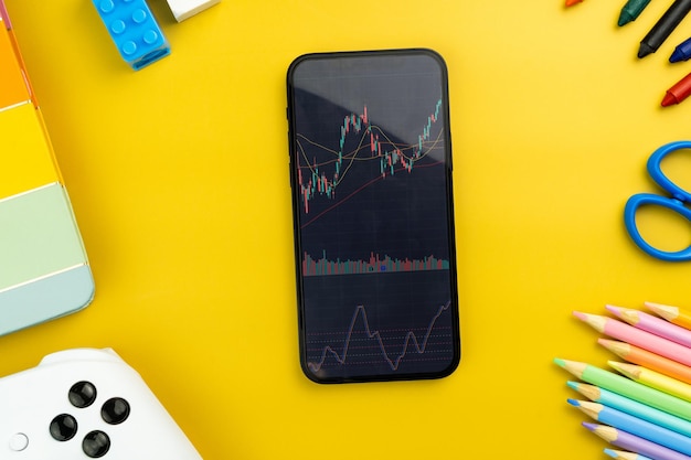 Financial stock market graph on the smartphone screen yellow\
background with school supplies children\'s accessories video game\
controller stock exchange