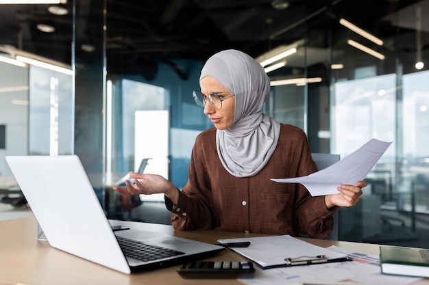 Financiële problemen bezorgden jonge moslim hijabi-zakenvrouw die op kantoor zit en aan laptop werkt en