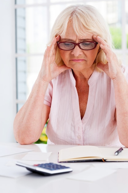 Financiële problemen. Gefrustreerde senior vrouw die het hoofd in handen houdt en naar de rekeningen kijkt die op tafel liggen