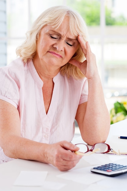 Financiële problemen. Gefrustreerde senior vrouw leunt haar hoofd bij de hand en houdt de ogen gesloten terwijl ze aan tafel zit met rekeningen en rekenmachine erop