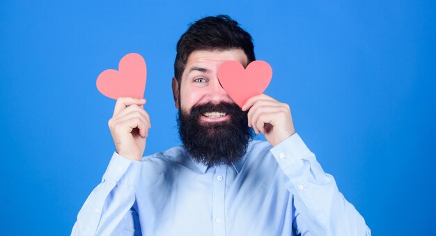 Find love with heart not eyes Bearded man hiding eye with red heart Hipster holding small hearts Sexy valentine man with heart shaped cards Happy valentines day The holiday of love and romance