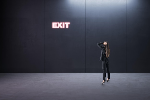 Trova la decisione giusta e il miglior concetto di via d'uscita con la vista posteriore della donna d'affari pensierosa nella stanza vuota guardando il segno di uscita al neon luminoso sulla parete scura