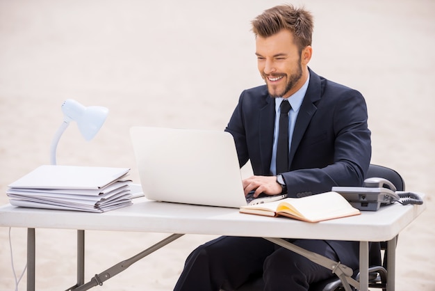 Finding a peaceful place to work. Cheerful young man in formalwear working on laptop while sitting at the table on sand