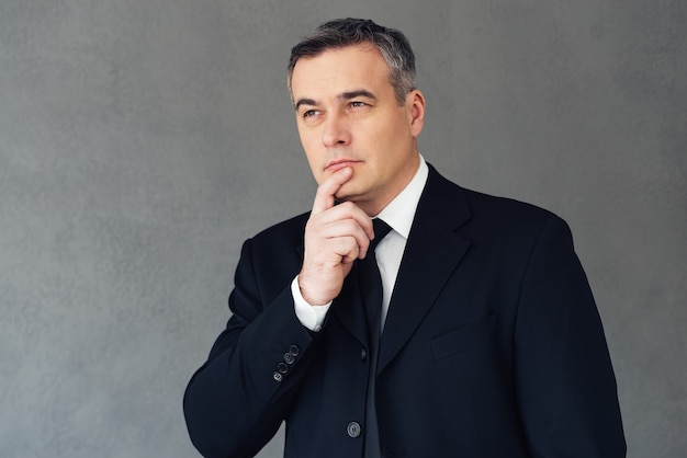 Finding perfect solution. Mature businessman holding hand on chin and looking thoughtful while standing against grey background