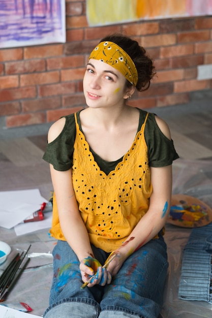 Fine art hobby. Portrait of young lady sitting on floor in studio, hands dirty with paint, holding brush.