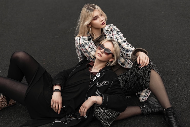 Photo サングラスをかけた黒い服を着た素敵な若い女性が、市松模様のブレザーを着た金髪のガールフレンドの隣のアスファルトに横たわっている
