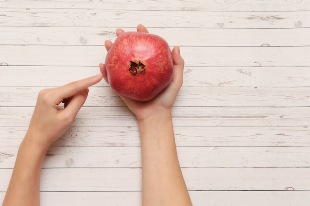 Finger points to whole unpeeled pomegranate in female's hand on white wooden table, top view