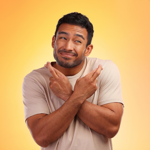 Fingers crossed man and model with a orange studio background with hand sign for luck smile hope and faith of a person with beard holding hands gesture for wish and optimism feeling hopeful