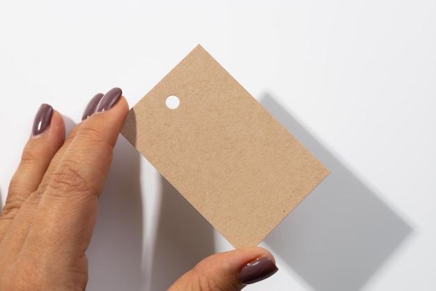 Fingers holding cardboard blank rectangular label tag with small hole in upper part for clothes in center with shadows falling on white background Tag mock up Copy space