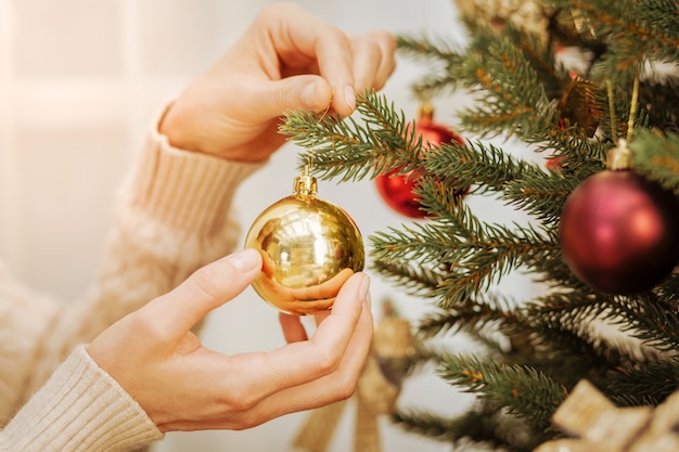 Finishing touches. Scaled up look on female hands holding a shiny golden ball while standing at a tree and decorating it for christmas.