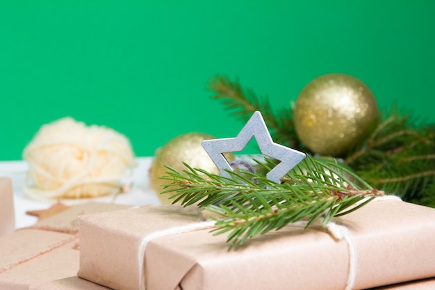 Fir branches, shiny gold balls for the Christmas tree and a gift box wrapped in kraft paper and decorated with a wooden star on a green background, eco christmas concept