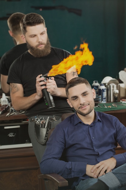 Fire haircut. Handsome man smiling sitting in a barber chair while his bearded barber spraying fire flames with a hairspray behind him