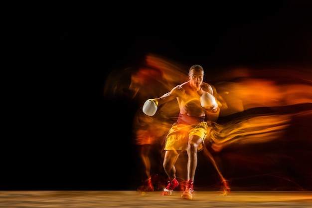 Photo in fire. professional boxer training isolated on black studio background in mixed light. man in gloves practicing in kicking and punching. healthy lifestyle, sport, workout, motion and action concept.