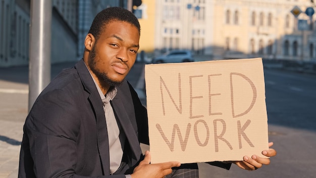 Fired sad unemployed businessman ethnic guy in business suit in city looking for job Millennial upset African American man male searching employment in crisis sitting outdoors with poster Need Work