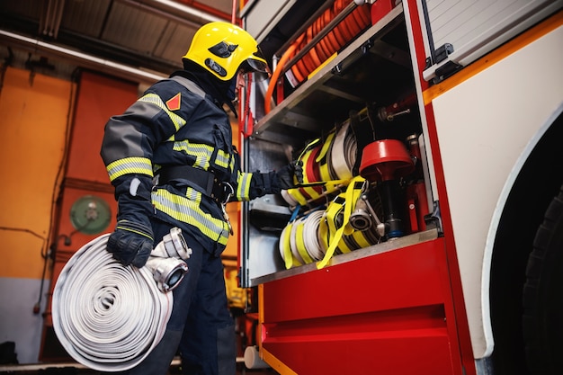 Photo firefighter in protective uniform with helmet on head checking on hoses before intervention while standing in fire station.