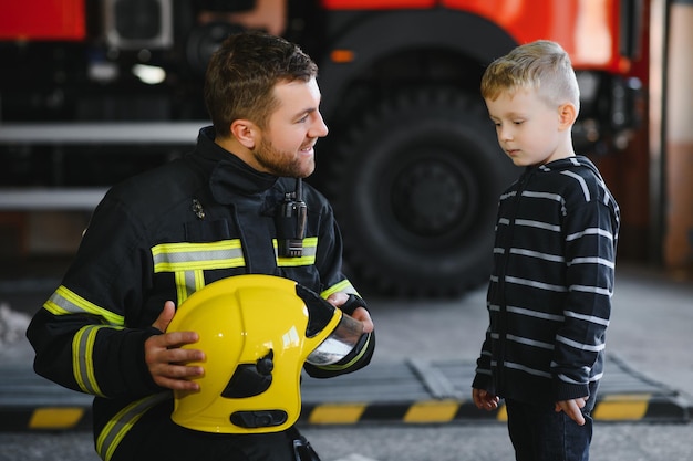 A firefighter take a little child boy to save him fire engine\
car on background fireman with kid in his arms protection\
concept