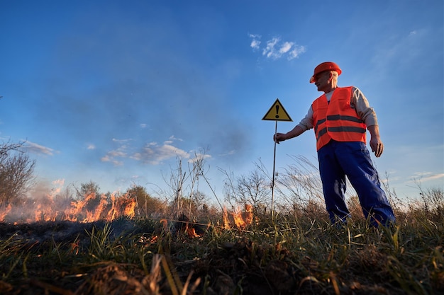 Fireman ecologist extinguishing fire in field in evening Man in orange vest and helmet near burning grass with smoke holding warning sign with exclamation mark Natural disaster concept