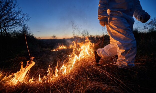 Fireman ecologist fighting fire in field at night Cropped view of man in protective radiation suit running through burning grass with smoke Natural disaster concept