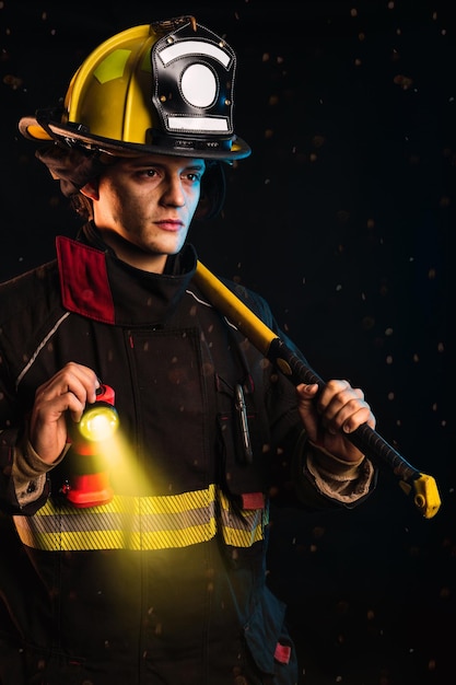 Vigile del fuoco in piedi con martello che indossa una speciale uniforme protettiva con torcia accesa sfondo nero notte e fuoco