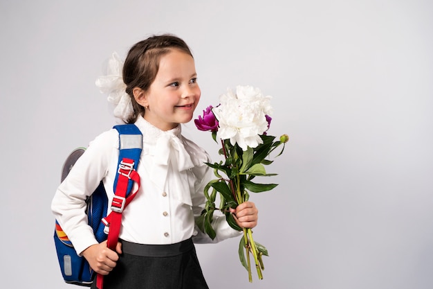 First grade school girl in a white shirt holding flowers and a backpack