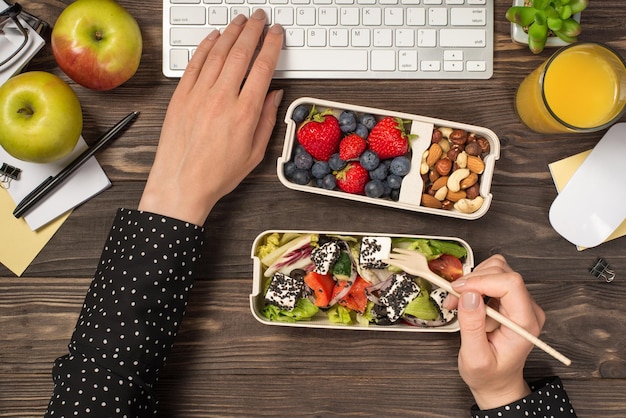 Фотография от первого лица, на которой женские руки едят здоровую пищу из двух коробок для завтрака и печатают на клавиатуре, мыши, стакан сока, яблоки, растение и канцелярские принадлежности на изолированном фоне темного деревянного стола