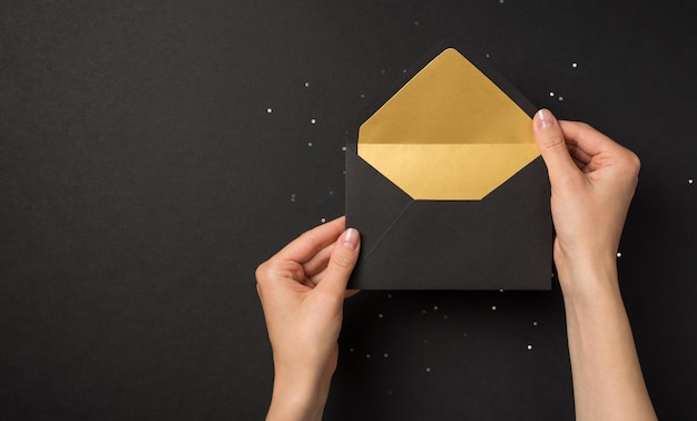 First person top view photo of female hands holding black and golden open envelope over sequins on isolated black background with empty space