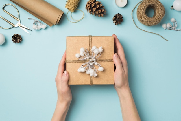 First person top view photo of female hands holding giftbox decorated with snow twig over balls scissors spools of twine roll of craft paper candle and cones on isolated pastel blue background