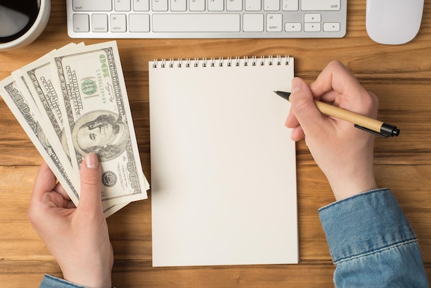 First person top view photo of female hands holding hundred dollars banknotes and pen over notepad cup of coffee keyboard mouse on isolated wooden desk background with blank space