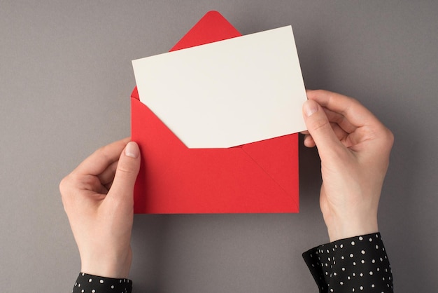 First person top view photo of female hands holding open red envelope with white card on isolated grey background with copyspace