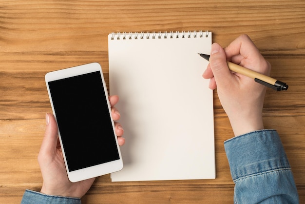 First person top view photo of female hands holding pen over planner and smartphone on isolated wooden table background with blank space