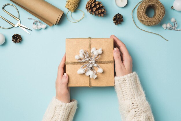 First person top view photo of female hands in knitted pullover holding giftbox with snow twig balls scissors spools of twine roll of craft paper candle and cones on isolated pastel blue background