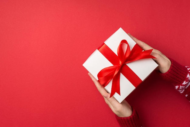 First person top view photo of female hands in red sweater holding big white giftbox with red ribbon bow on isolated red background with copyspace