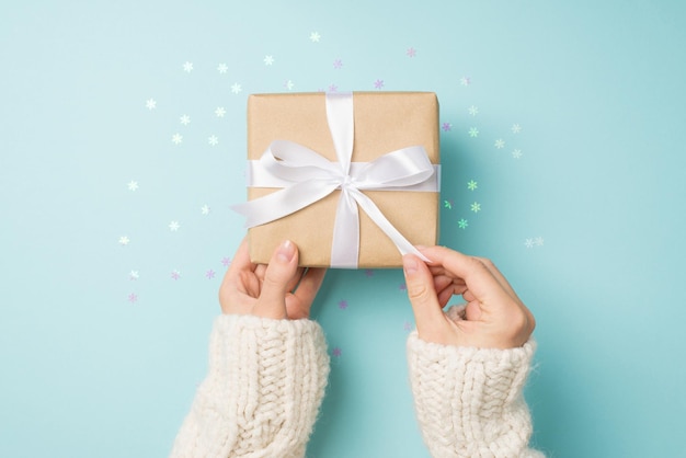 First person top view photo of female hands in white pullover opening craft paper giftbox with white ribbon bow over shiny snowflakes confetti on isolated pastel blue background