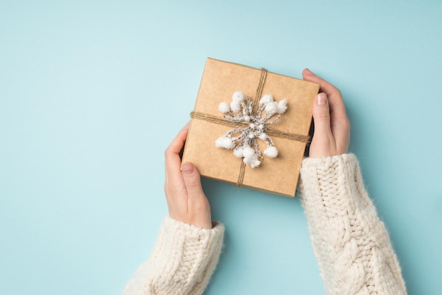 Foto in prima persona vista dall'alto di mani femminili in maglione bianco che dimostrano una confezione regalo di carta kraft decorata con ramoscello di neve e spago su sfondo blu pastello isolato