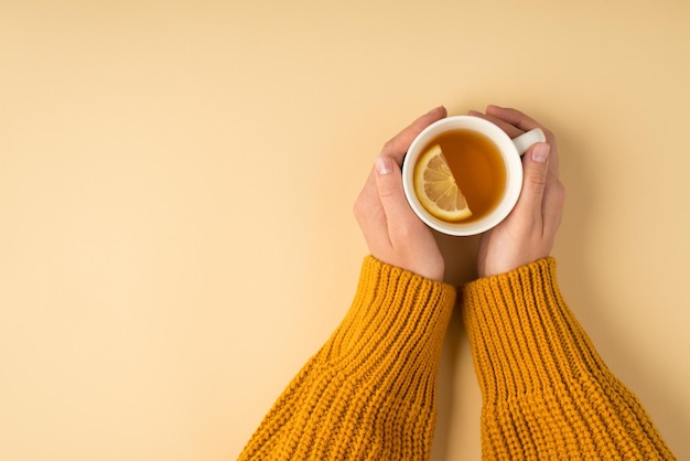 Foto in prima persona vista dall'alto di mani femminili in maglione giallo che tengono una tazza di tè bianca con fetta di limone su sfondo arancione pastello isolato con copyspace