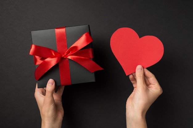 Foto in prima persona vista dall'alto delle mani della ragazza che tengono cuore di carta rossa e confezione regalo nera con fiocco di nastro rosso su sfondo nero isolato con spazio vuoto