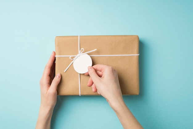 First person top view photo of girl's hands holding pricetag on craft paper giftbox with small white bow on isolated pastel blue background with blank space