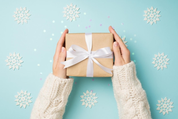 First person top view photo of girl\'s hands in white sweater\
holding craft paper giftbox with white ribbon bow over big\
decorative snowflakes and shiny sequins on isolated pastel blue\
background