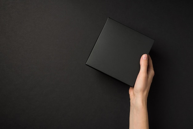 First person top view photo of hand holding black unwrapped giftbox on isolated black background with empty space