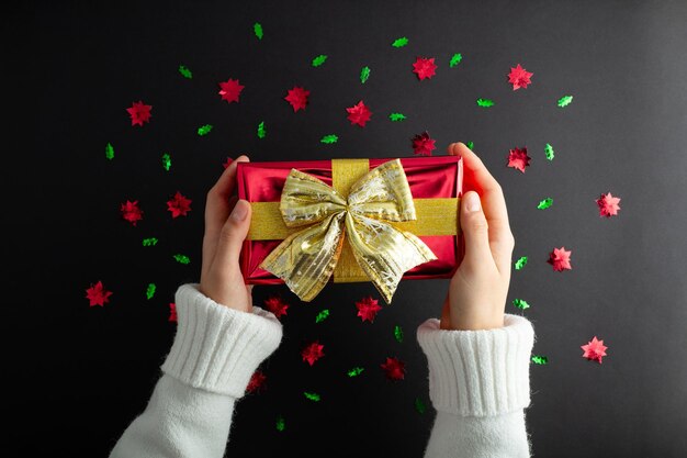 First person top view photo of hand holding redgiftbox with golden satin ribbon bow over shiny red and green sequins on isolated black background with empty spacexA