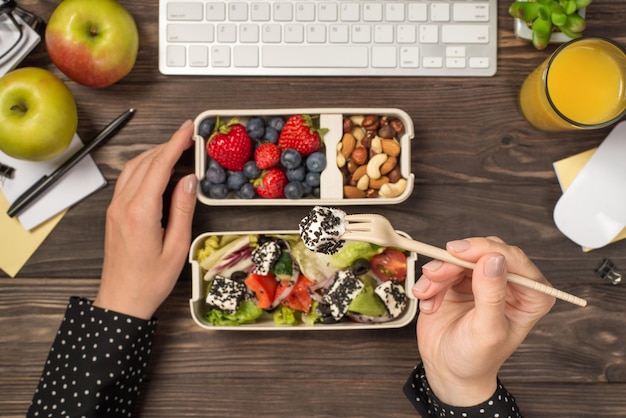 First person top view photo of hand holding tofu on fork over two lunchboxes with salad berries nuts apples glass of juice plant stationery keyboard and mouse on isolated dark wooden table background