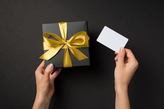 Photo first person top view photo of hands holding black giftbox with golden satin ribbon bow and white plastic card on isolated black background with blank space