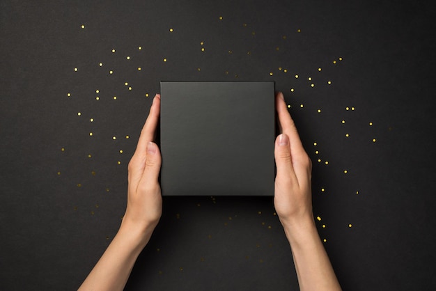 First person top view photo of hands holding black unwrapped giftbox and golden sequins on isolated black background with blank space