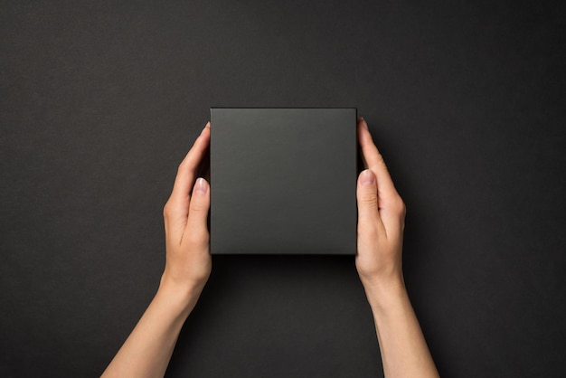 First person top view photo of hands holding black unwrapped giftbox on isolated black background with blank space