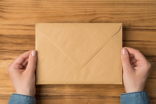 First person top view photo of hands holding closed craft paper\
envelope on isolated wooden table background with copyspace