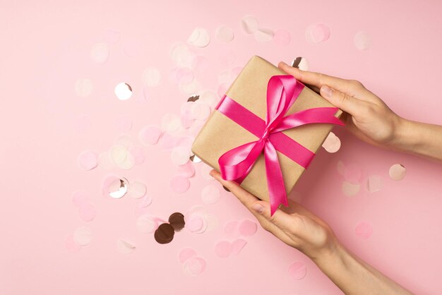 First person top view photo of hands holding craft paper giftbox with pink ribbon bow over large shiny sequins on isolated pastel pink background with empty space